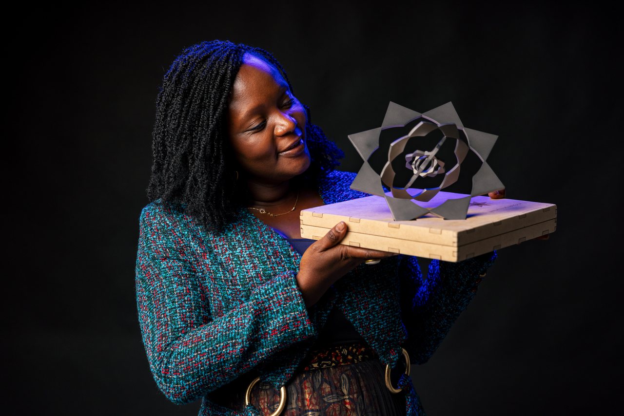 Chef Binta, lit against a dark backdrop, holds an open award or certificate in both hands.