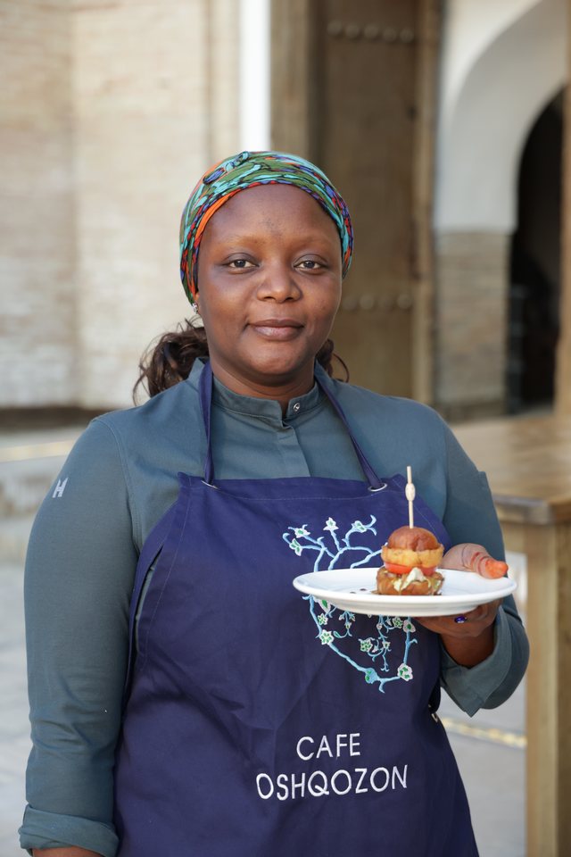 Chef Binta wearing a “Café Oshqozon” apron, holding a plate with a pastry, standing before an ornate archway.