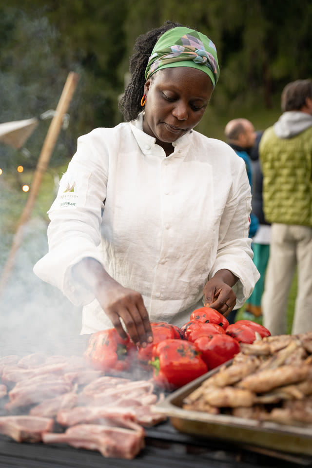 Chef Binta, in a green headscarf, grilling meat and tomatoes over an open outdoor fire.