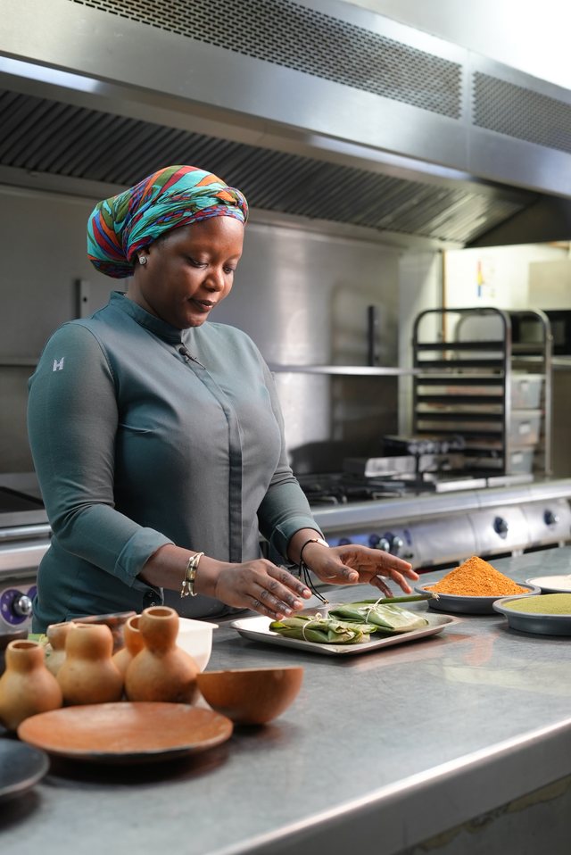 Chef Binta in a professional stainless-steel kitchen, plating a dish, wearing a colorful headwrap.