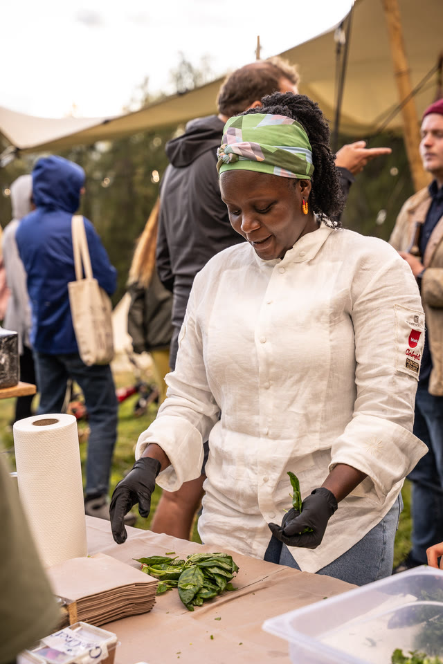 Chef Binta, in a green headscarf, preparing ingredients at an outdoor table beside a tent.