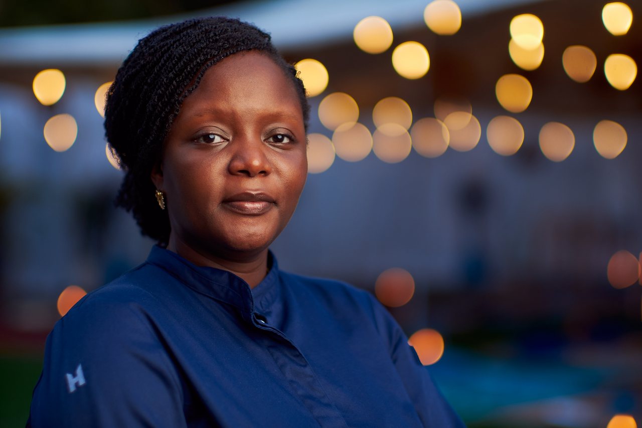 Chef Binta smiling, lit warmly against a background of blurred string lights at night.