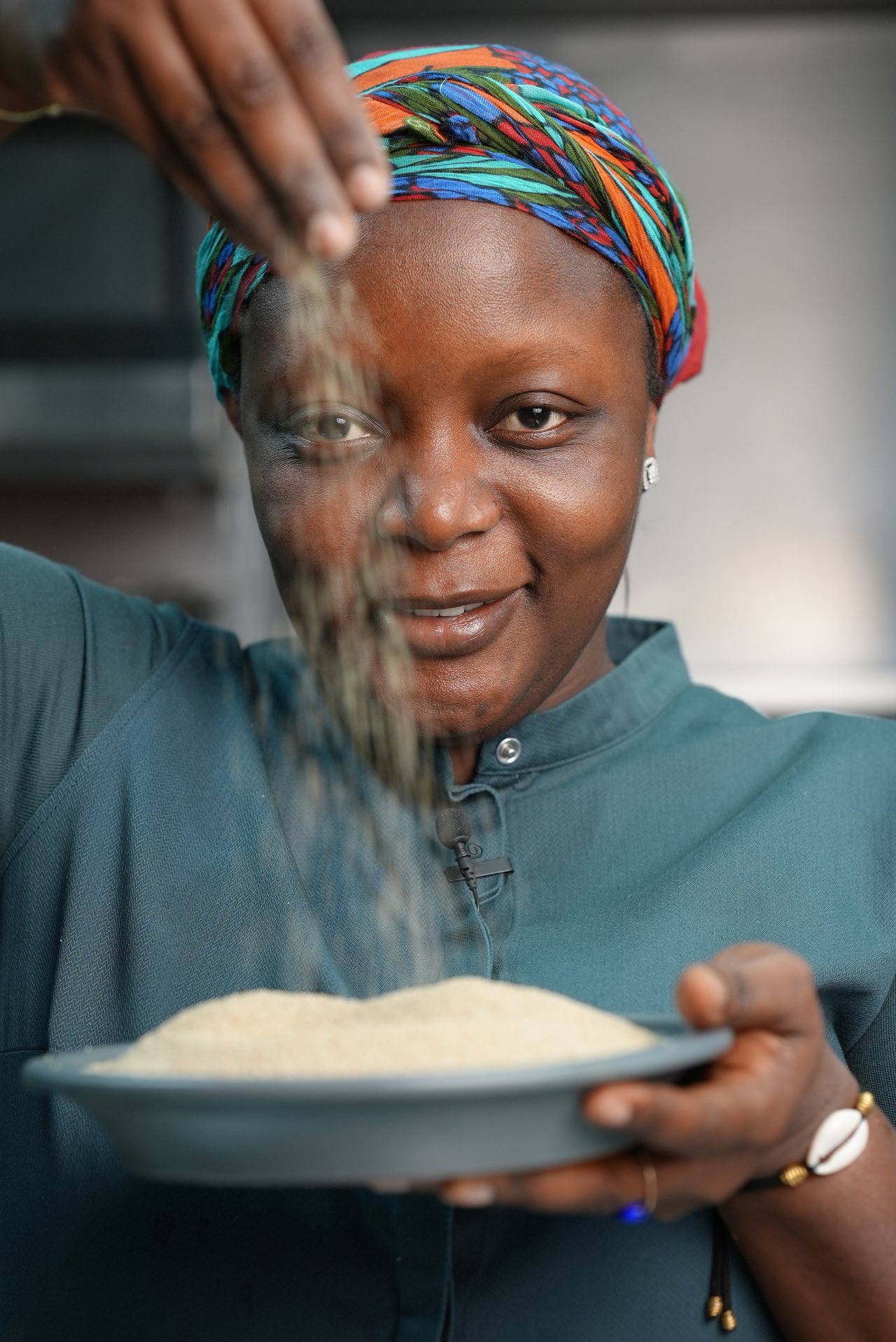 Close on Chef Binta's face and hands as she sprinkles seasoning onto a plate, warm light, colorful headwrap.