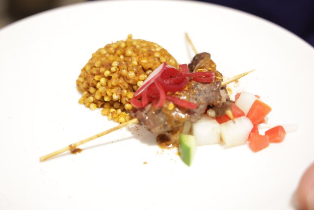 Looking down at a mound of yellow grain topped with a skewer and diced vegetables, on a white plate.