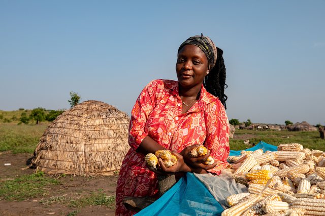 A woman in a red patterned dress sorts produce on the ground outside a thatched hut.