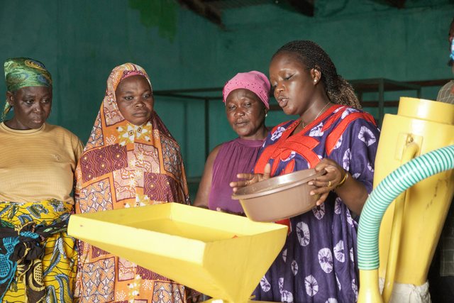 A group of women in colorful patterned dress stand around a yellow mechanical mill.
