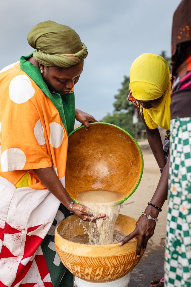 Two women in orange and yellow pour liquid from a calabash into a carved wooden mortar, washing the grain.