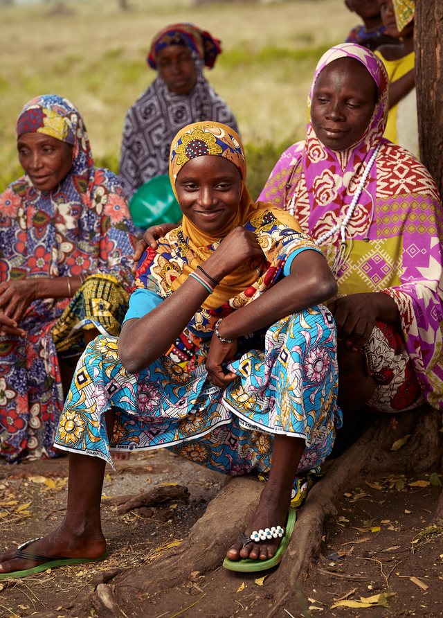 A group of young women and girls in colorful patterned dress sit together outdoors, smiling.