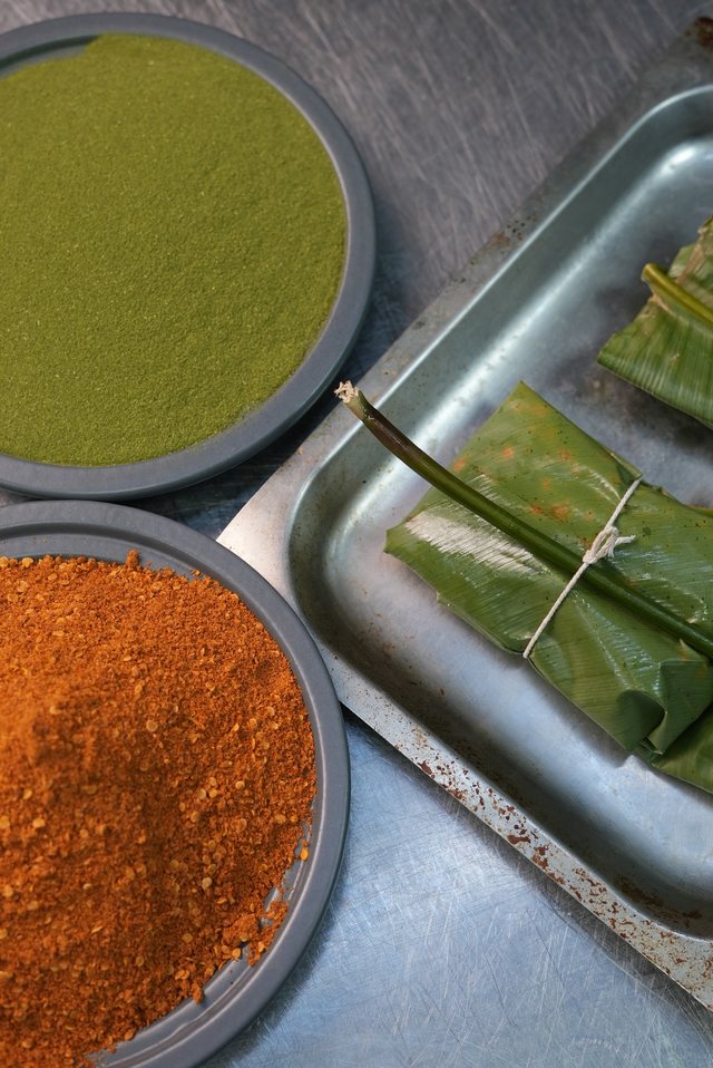 An overhead styled shot of ingredients: a bowl of green powder, a bowl of red spice, and leaf-wrapped bundles on a metal tray.
