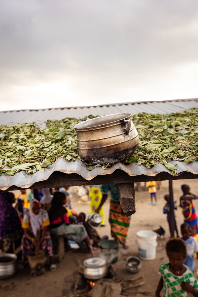Overhead view of a large pot on an outdoor fire, greens drying on a rack above, market activity below.