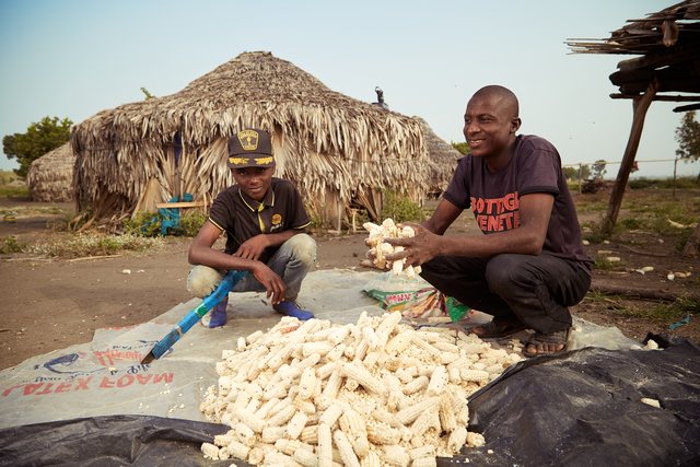 Two men crouched on a mat outdoors, processing roots or produce in front of a thatched hut.