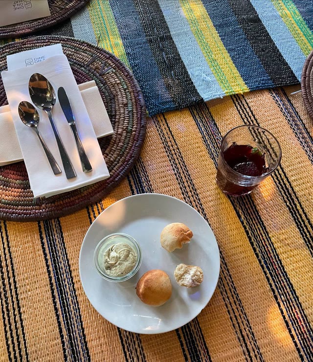 Overhead view of a place setting on a woven mat: cutlery, a small plate of bread and dip, and a glass of wine.