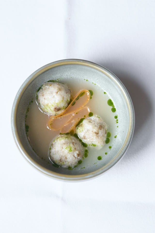 Overhead view of a white bowl holding three pale dumplings in a light broth, garnished with herb oil.