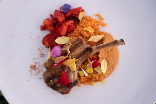 A lamb chop plated with a colorful garnish of fruit, herbs and spice, against a pale marble table.