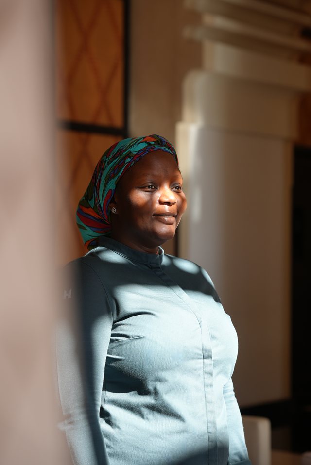 A woman in a teal top standing beside a stone or wooden pillar, soft indoor light.