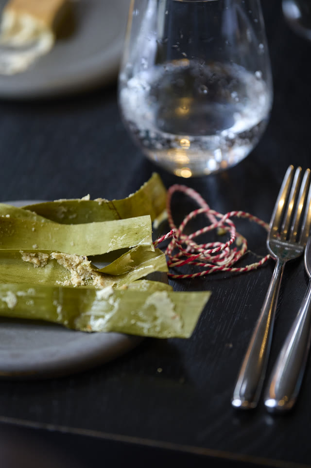 A place setting close up: a wine glass, a leaf-wrapped parcel tied with string, and cutlery on a dark table.