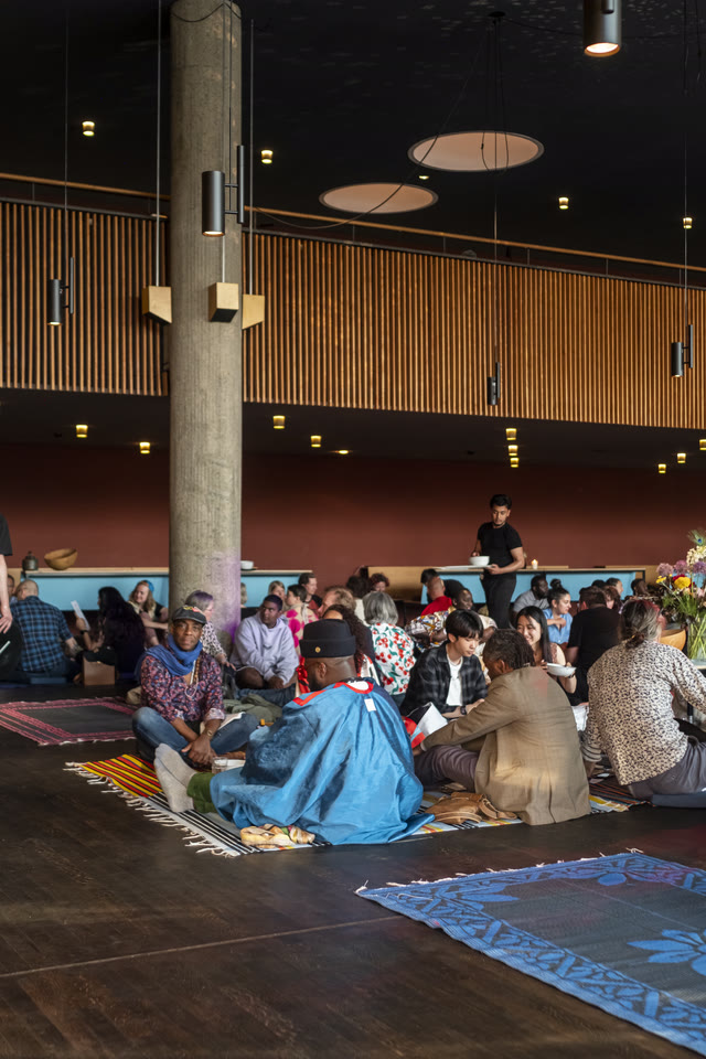 A wide, low room with concrete columns, patterned rugs on the floor, and guests seated for dinner.