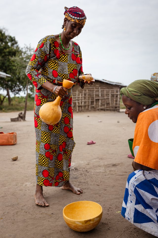 A woman in a red patterned dress pours liquid from a gourd, a child standing nearby.
