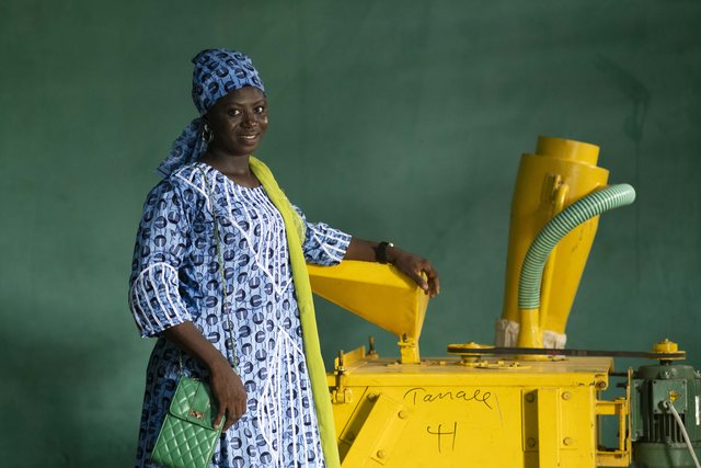 A woman in blue stands beside a yellow mechanical grain-milling machine outside a thatched structure.