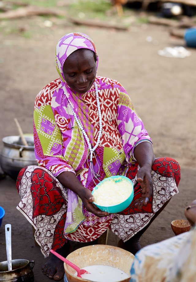 A woman in a vivid purple and pink patterned dress sits holding a white bowl of dough or dairy.